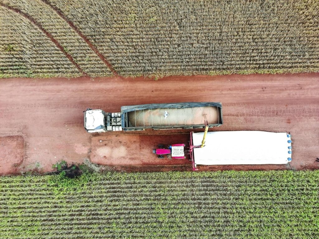 An aerial view captures larges trucks being loaded with harvested grain in a vast agricultural field. The image highlights the process of grain harvesting and transportation, showcasing modern farming techniques and machinery. The scene is set against a backdrop of a mature crop field.