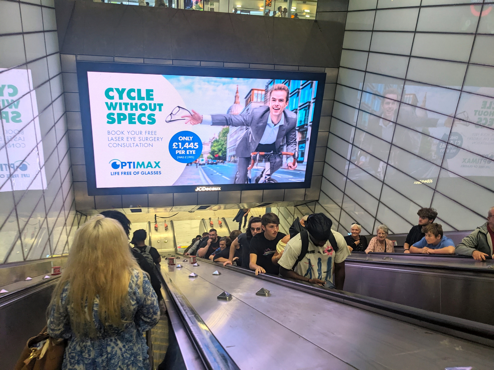 Commuters ride a busy escalator past a large digital advertising billboard in a London Tube station