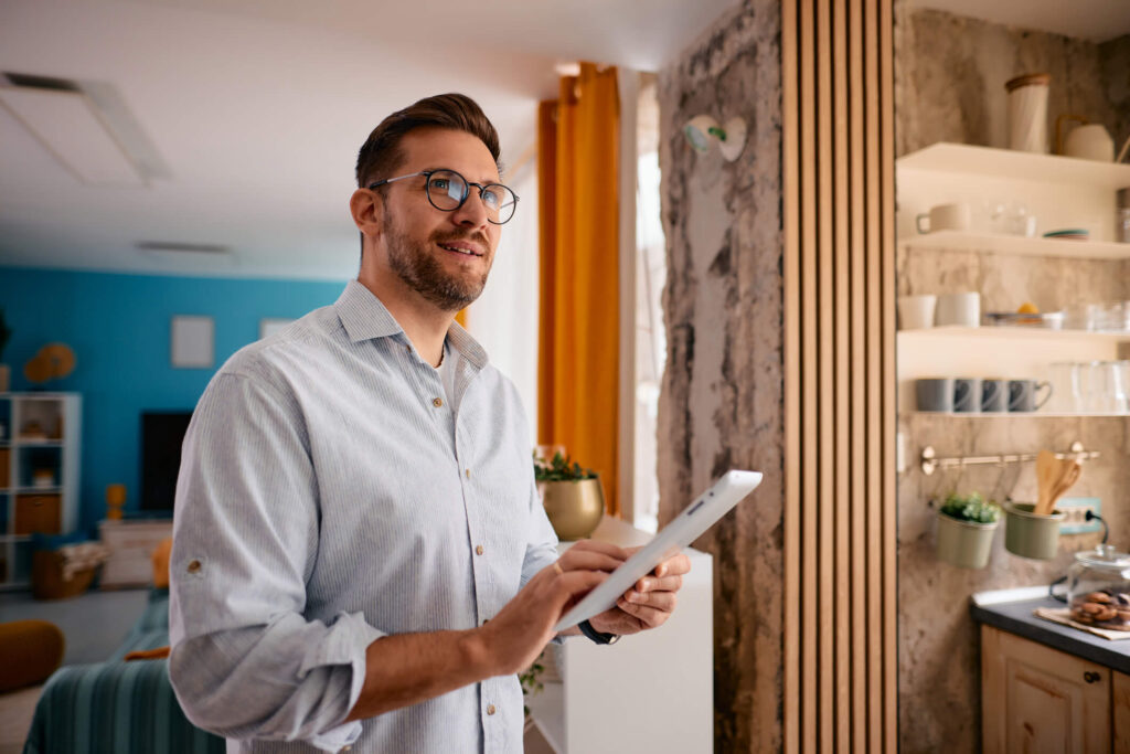 Mid adult businessman standing confidently in a modern apartment, using a digital tablet and gazing thoughtfully out the window while working from home, surrounded by natural light