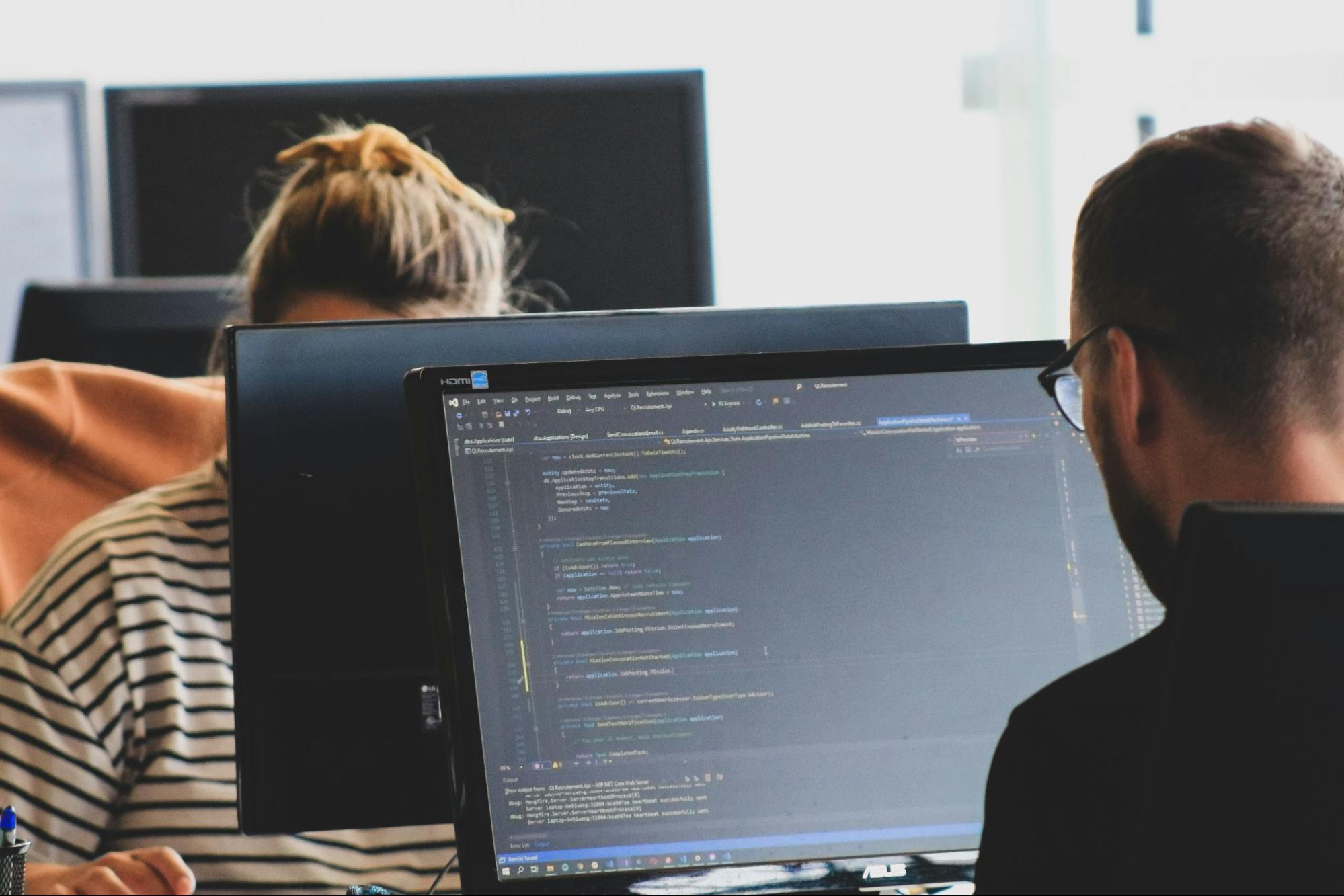 Software developers reviewing code on desktop monitors in an office, representing code review and maintenance practices used to reduce technical debt