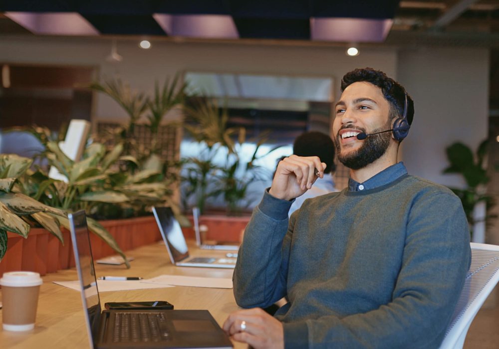 Young consultant with beard wearing headset smiling cheerfully and listening to customer in call center